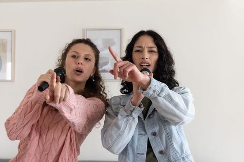 Two friends singing duet pointing forward with microphones in cozy living room