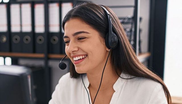 Smiling female customer service agent wearing headset at office desk with monitor and binders