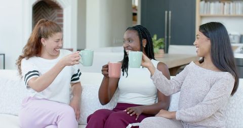 Three Friends Clinking Pastel Mugs on Cozy Sofa in Bright Living Room Casual Loungewear