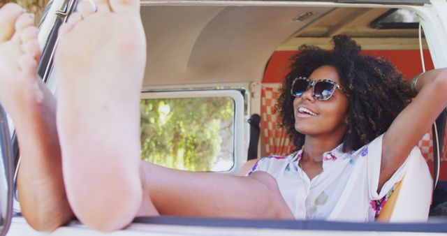 A young African American woman relaxes in a vintage van, her feet up and sunglasses on, with copy space. Her carefree pose and bright smile evoke a sense of summer adventure and freedom.