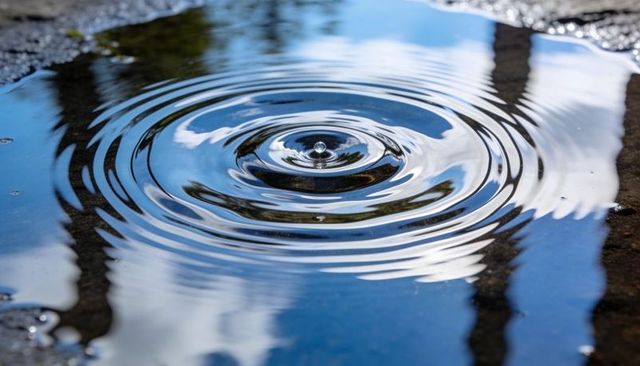 Single water droplet creating concentric ripples reflecting blue sky on pavement puddle