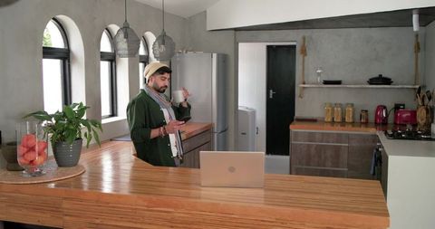 Indian man in beanie standing at wooden kitchen island holding mug and checking smartphone