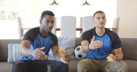 Friends cheering and reacting to soccer match on couch eating pizza in team shirts
