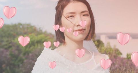 Bride wearing short veil standing in garden with floating pink hearts, pastel romance