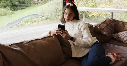 Young woman relaxing on sofa using smartphone wearing festive reindeer headband