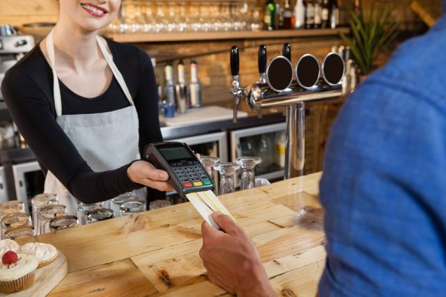 Customer making payment with credit card at cafe counter. Barista holding POS terminal while customer completes transaction. Ideal for illustrating modern payment methods, small business operations, and customer service in retail or hospitality settings.