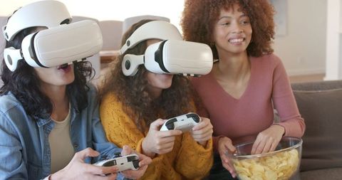 Women playing virtual reality games on couch sharing snacks and laughing together