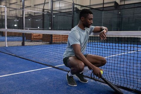 African american athlete crouching by net on indoor padel court checking smartwatch holding ball