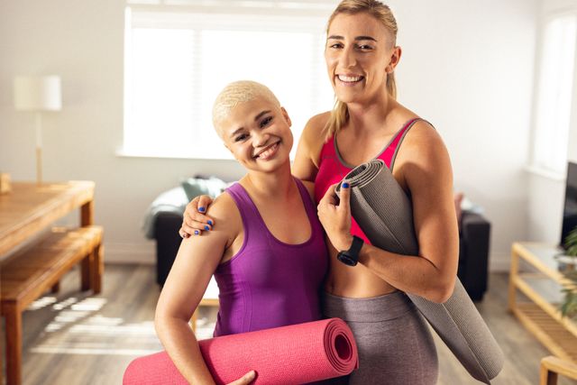 Two cheerful young women, a lesbian couple, are holding yoga mats and smiling at home. This image can be used for promoting healthy living, fitness routines, LGBTQ representation, and lifestyle blogs. It is ideal for content related to exercise, home workouts, and relationship bonding.