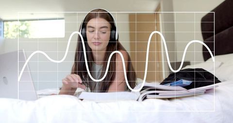 Woman listening with headphones studying on bed with laptop and notebooks, waveform overlay