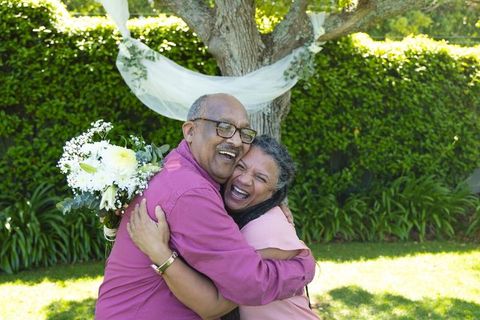 Mature couple embracing and laughing in sunlit garden with bridal bouquet and greenery