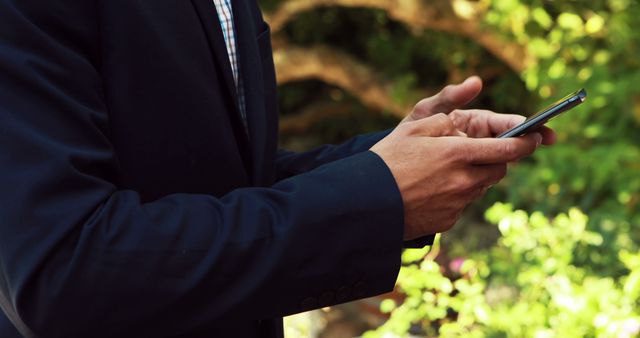 A businessman in a dark suit uses a smartphone outdoors in a lush garden, suggesting themes of business communication, technology, and connectivity. This image can be used for articles on remote working, business communication, technology in business, or for advertising business-related mobile apps and services.
