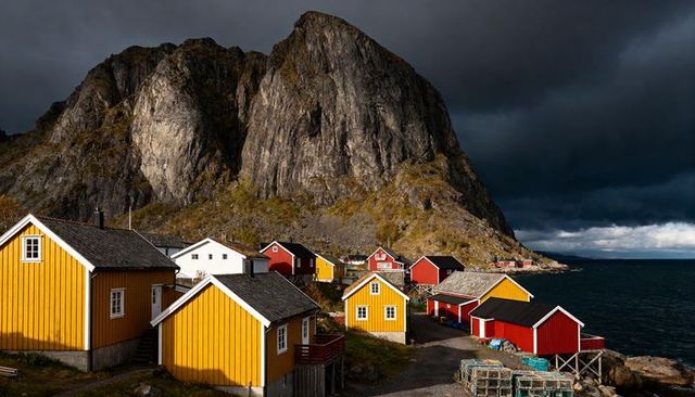 Dramatic norwegian fishing village with yellow and red cabins beneath towering rock and stormy sky