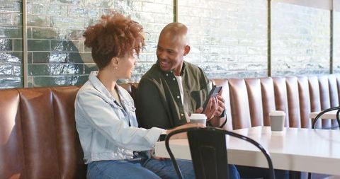 African american couple sharing smartphone and coffee while sitting on cafe booth, smiling