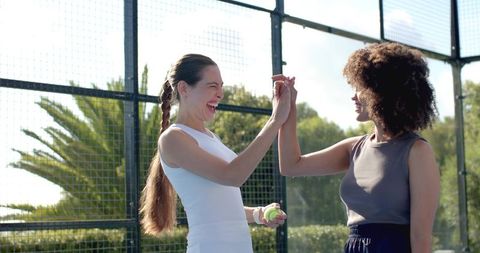 Female friends high-fiving on outdoor tennis court while holding lime tennis balls