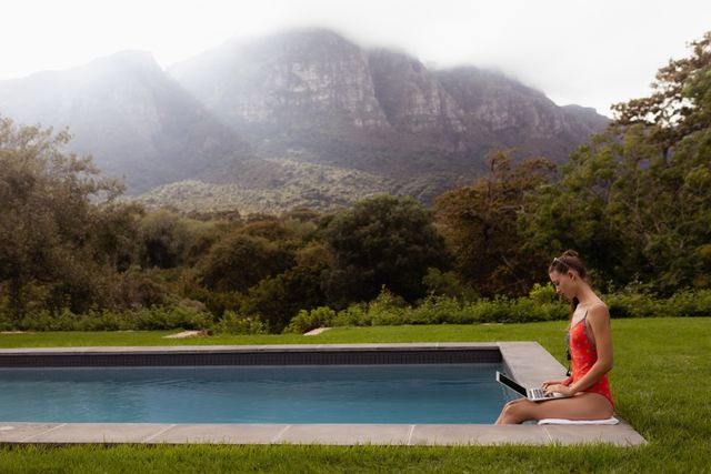 This image depicts a woman in swimwear sitting by the poolside, using a laptop with a scenic mountain view in the background. Ideal for illustrating concepts of remote work, relaxation, summer activities, and outdoor leisure. Perfect for use in travel blogs, lifestyle articles, and advertisements promoting work-life balance or vacation destinations.