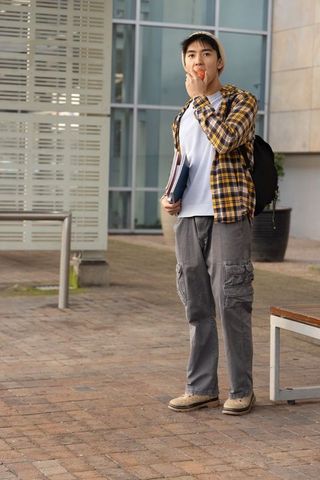Asian teen student standing on campus eating apple holding notebooks and backpack