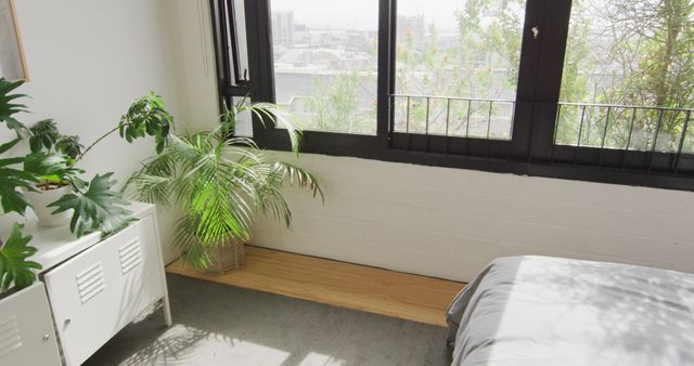A modern, urban bedroom featuring a large window that allows ample sunlight to fill the space. Several indoor plants create a tranquil and inviting environment. The minimalist setting complemented by clean lines provides a sense of peace and coziness. This image can be used to illustrate modern home decor, urban living, or creating green spaces indoors.