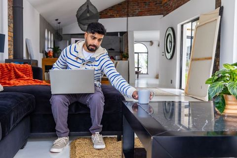 Man working on laptop in modern loft living room reaching for coffee mug and relaxing
