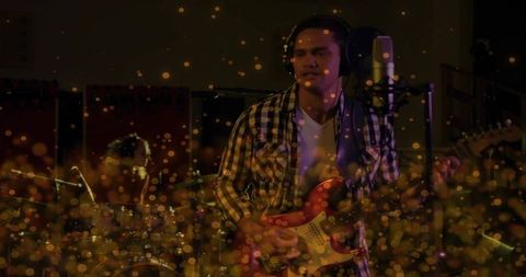 Recording male guitarist with headset in plaid shirt holding red electric guitar amid bokeh lights