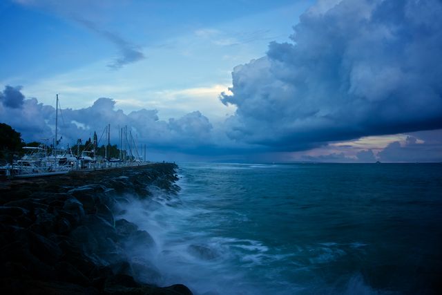 Dramatic scene of a seaside marina with large storm clouds forming at dusk. Boats are docked along the rocky shoreline, as waves crash against the rocks. Useful for illustrating weather phenomena, nature's power, or serene coastal life.