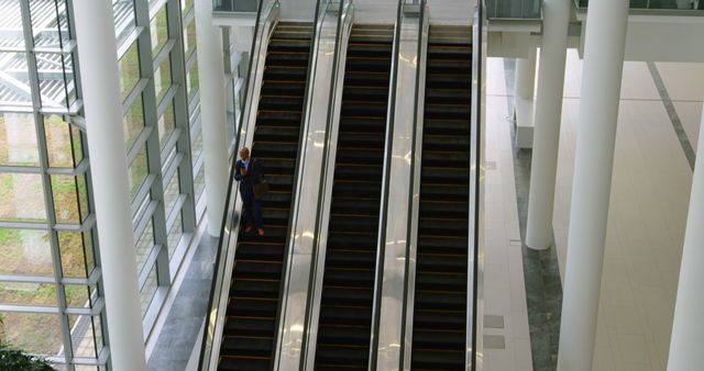 Business professional standing on escalator in modern office building. Image conveys themes of professional life, upward mobility, and corporate workspace. Can be used for website banners, corporate presentations, and business-related marketing materials.