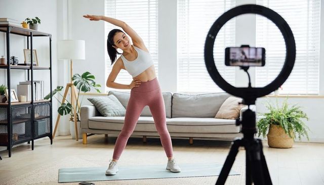 Female fitness instructor streaming live home workout with ring light and smartphone