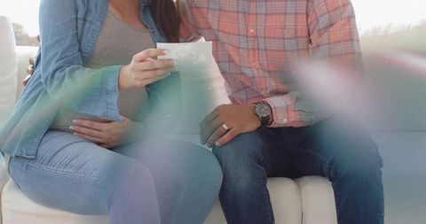 Expecting couple holding ultrasound printout on cozy sofa, wedding ring and watch closeup