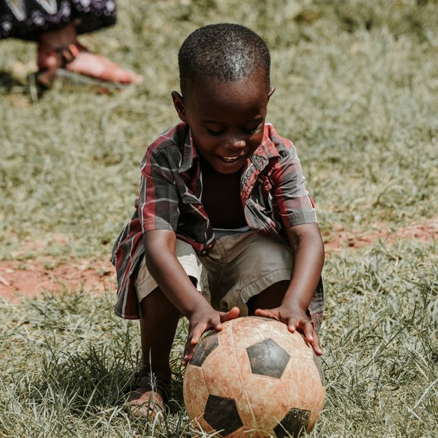 Young African boy is playing with soccer ball outdoors on a sunny day. Wearing a plaid shirt and casual shorts, he is handling soccer ball on grass field. This can be used for themes of childhood, playtime, sports activities, outdoor fun, health and fitness, sunny weather, and cultural diversity.