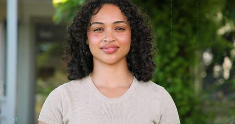Smiling woman wearing beige knit top posing at entrance with lush green background
