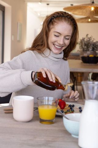 Woman enjoying cozy breakfast pouring syrup on pancakes at home