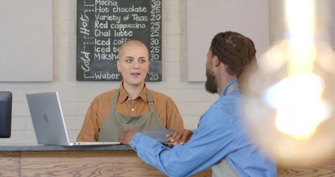 Two baristas collaborating behind wooden counter with laptop and chalkboard menu, cozy vibe