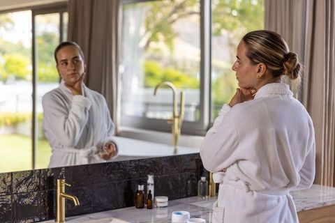 Woman inspecting skin in mirror at bath vanity wearing white robe for skincare routine