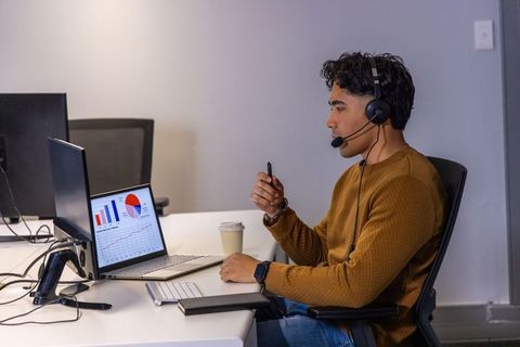 Indian man in 20s analyzing laptop charts wearing headset at minimalist office desk