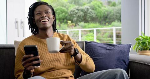 African American Man Relaxing on Sofa Drinking Coffee and Using Smartphone