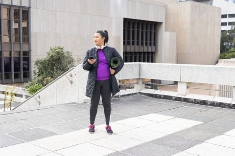 Mature woman standing on urban rooftop holding yoga mat and smartphone in quilted coat