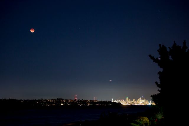 Lunar eclipse casting reddish glow in night sky above city skyline illuminated by bright city lights. Ideal for themes related to astronomy, urban night photography, celestial observations, and cityscapes.