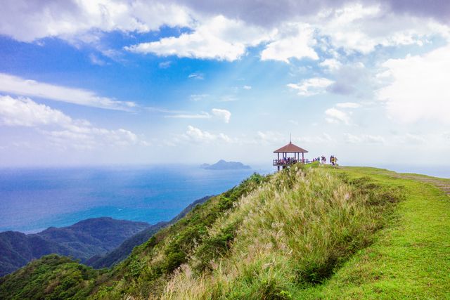 Panoramic view highlighting a serene gazebo on a mountain ridge overlooking the ocean in the distance. Lush green landscape meets vibrant blue sky with fluffy clouds, creating a peaceful and idyllic scene. Ideal for travel brochures, outdoor adventure advertisements, nature calendars, and backgrounds for websites focusing on travel and nature.