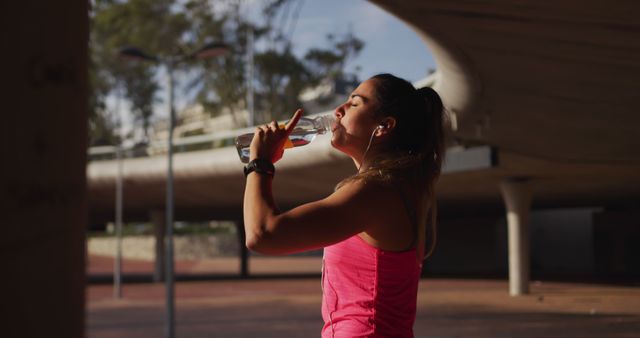 A young woman, wearing a pink workout top, is drinking water while standing under a shaded structure. She appears to be recovering after an outdoor workout or run. This image can be used to promote healthy lifestyles, fitness routines, hydration importance, and outdoor exercise activities.