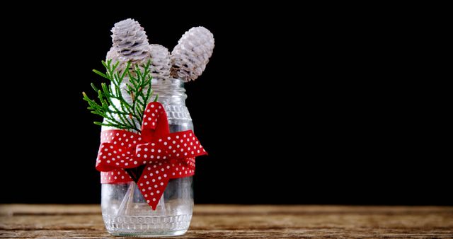 A clear glass jar adorned with a red polka dot ribbon contains pine cones and greenery, with copy space. Its rustic charm and festive decoration suggest a simple, yet elegant holiday centerpiece or home decor.