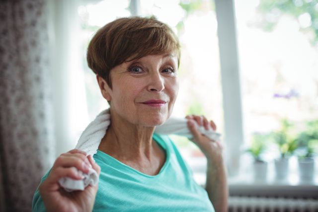 Senior woman holding towel around neck, smiling at home. Ideal for use in articles about healthy aging, senior fitness, relaxation, and home life. Can be used in advertisements for wellness products, senior care services, or lifestyle blogs.