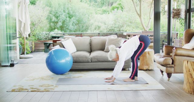 A senior woman practices yoga in her living room, using a yoga mat and exercise ball. The room features large windows with a view of greenery, giving a sense of calm and tranquility. This stock photo is ideal for promoting fitness, healthy living, aging gracefully, and home workout resources.