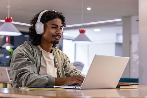 Indian man wearing white headphones typing on laptop at modern cafe coworking table