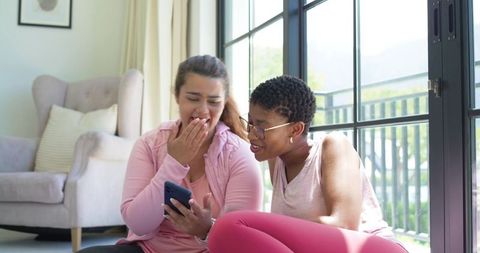 African american friends laughing and sharing phone while sitting on living room floor