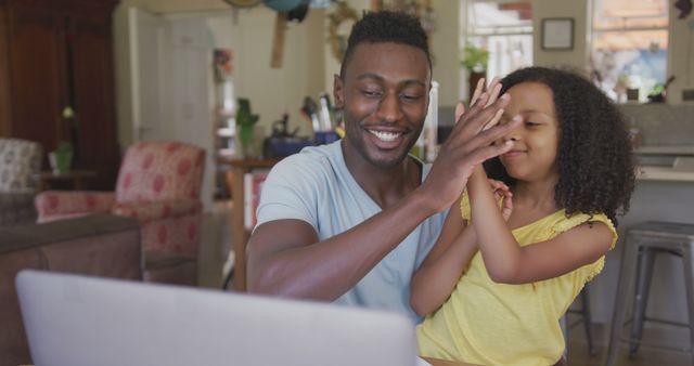 Happy african american father and daughter sitting at table using laptop and high fiving. Fatherhood, childhood, fun, togetherness and domestic life.