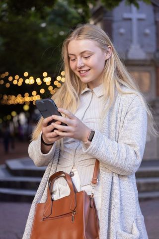 Young woman smiling and texting on smartphone in urban plaza at dusk with leather tote