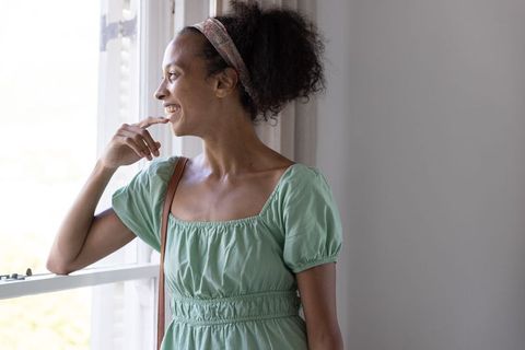 African american woman smiling and gazing out open window in sunlit home interior