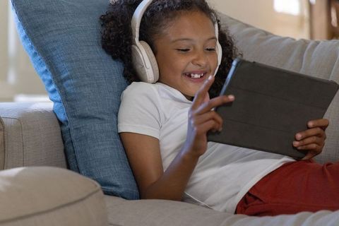 African american child reclining on sofa wearing headphones and using tablet, smiling