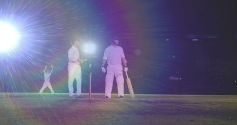 Night cricket batsman standing at crease under bright floodlights with celebrating fielder