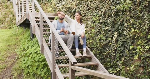 Diverse couple sitting on wooden stairs pointing and holding reusable bottles by ivy wall